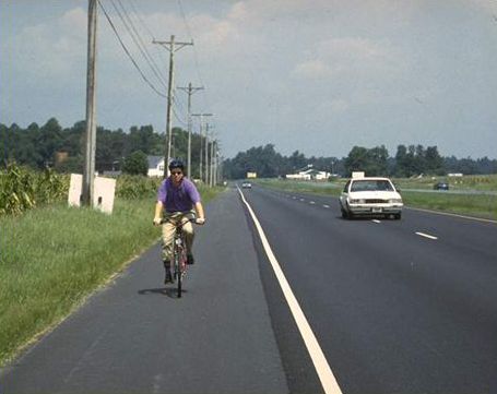 The picture shows a 4-lane road, two lanes in both directions, passing cornfields. The road has a paved 9’ shoulder, and a bicyclist is riding towards the camera.