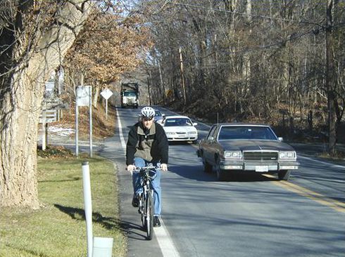 The picture shows a cyclist riding on the 1 foot paved shoulder of a busy two lane suburban road. There is a truck approaching the cyclist from behind and in order for the cars to pass the cyclist, they must cross the solid yellow center line.