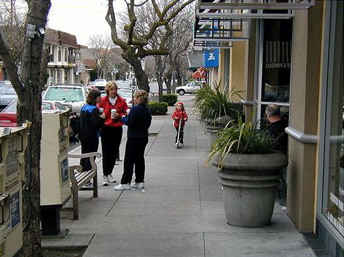 Picture shows an 8-12’ sidewalk in front of stores. There are trees, large flower pots, newspaper boxes and park benches, narrowing the walking area. There is a child on a scooter and three women holding coffee cups and chatting outside one of the shops.