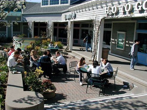 The picture shows a sunken patio area with café tables in what looks like a new shopping center. Parking does not come right up to the doors of the shops. There is a covered pergola area enclosing the patio, and the shop entrances are under this covered area. The picture shows numerous groups of people sitting on a wall separation of the patio, and others seated around tables.