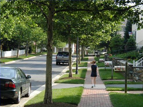 Picture is of a lovely suburban neighborhood with a tree lined street, well tended yards and a wide brick paved sidewalk in front of the houses. There are no garages visible. There are a couple cars parked along the shaded street and a child is walking on the sidewalk in a direction away from the camera.