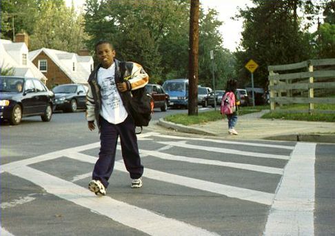 Picture shows a boy walking to school. He is crossing in a wide crosswalk and carrying a book bag. Cars wait for him to pass.