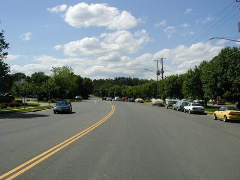 The picture shows an extremely wide 2-lane road with a car driving toward the viewer. The street looks like it is wide enough to be a four lane road, but it is striped only with a centerline. The road looks like it is a place where speeding is a problem.