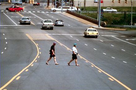 Second picture shows a wide urban arterial street in a strip shopping area. There are three lanes of traffic each direction with a center turning lane. The road is very wide, and two men are shown crossing the street, striding across the center turn lane.