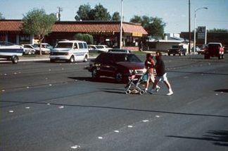 First picture is of street in a suburban shopping area with fast-food restaurants and six lanes of traffic. There are two women with a baby stroller crossing mid-block. A car is stopped mid-turn waiting for them to cross.