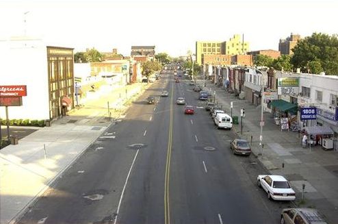 Picture shows an aging main street. There are wide sidewalks in front of shops. There are four wide traffic lanes as well as parking on both sides of the street. There are no trees or landscaping.