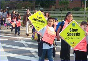 Three pictures are shown. All the pictures are of smiling children crossing at crosswalks. This picture shows children holding signs that read 'Safe Sidewalks' and 'Slow Speeds.'