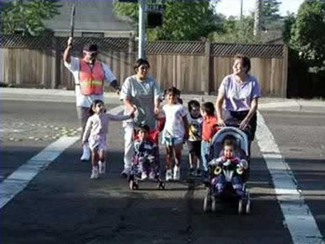 Picture shows two adults with six children crossing the street in a wide crosswalk. An adult crossing guard is assisting the group by holding a stop sign to stop traffic.
