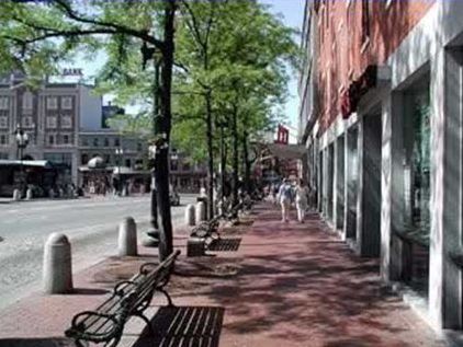 Picture shows a sidewalk between and street and store fronts, and benches, street trees and streetside bollards are also in this sidewalk space. 