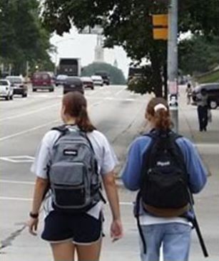 Picture shows two young women of high school age walking along a sidewalk. The young women both have backpacks strapped on their shoulders.