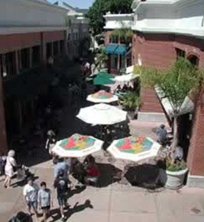 This picture shows a pedestrian plaza with dining tables covered by umbrellas. Several people are dining at the tables, and other people are walking through the plaza.