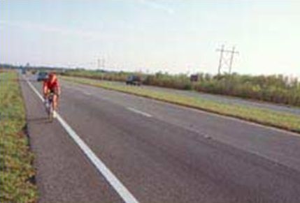 This picture shows a rural roadway that has a paved shoulder. A bicyclists is riding on the paved shoulder, with a few vehicles approaching in the far distance.
