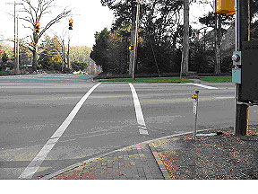 Figure 11. Photograph.  Crosswalk view of pedestrian example intersection. This is one of three photographs of the same intersection. This picture, from the street level, shows the signalized intersection of two roads, a four-lane main road and a two-lane cross street in a predominantly residential area. There is a marked, signalized crosswalk across the main road from one of the corners of the intersection.