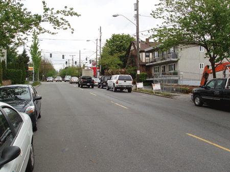 Figure 4. Photo. Fremont Street in the before period. The photo shows Fremont Street in Seattle, WA, in the before sharrow condition. There are two travel lanes, one in each direction, parked cars on both sides of the street, and a 3.6 percent grade on the site where the sharrows would be placed. The photo is shot from the left lane with the parked cars on the left side facing forward. The street is lined with buildings and houses.