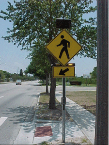 Figure 1. Photo. RRFB with two forward-facing LED flashers and a side-mounted LED flasher. A pole mounted at the side of a roadway near a crosswalk bears the W11-2 pedestrian warning sign, which shows a silhouette of a person walking to the left. The sign is a yellow diamond with a black border, and the silhouette is black. Below the sign, there is a rectangular rapid-flashing beacon (RRFB) with two yellow lights, and the light on the right is activated. Below the sign, there is a rectangular yellow sign with a black border and a black arrow pointing diagonally downward to the left toward the crosswalk. Above the W11-2 sign, there is a photovoltaic panel.