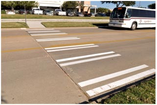 This photo shows a bus approaching a bar pair crosswalk. The crosswalk markings consist of several sets of two parallel white vertical bars across the width of the street.