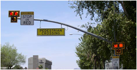 This photo shows a High intensity Activated crossWalK (HAWK) beacon on a street. Beacon heads consisting of two red lenses above a single yellow lens are located on both the masthead and a roadside pole. The red lenses are illuminated. Signs reading “CROSSWALK STOP ON RED” are positioned near the beacons, and a sign reading “PEDESTRIAN CROSSING” hangs from the masthead.