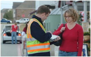 An experimenter gives an intercept survey to a participant outside a local grocery store. The experimenter is wearing a reflective safety vest and recording the participant's answers on an electronic device.