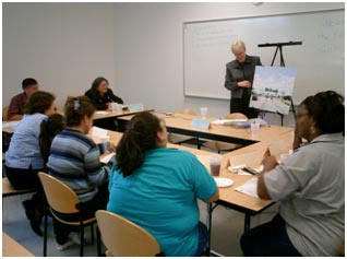 An experimenter conducts a focus group with six study participants. The participants are positioned around a table facing the experimenter, who is showing them an enlarged photo.