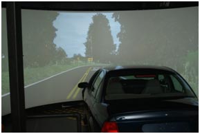A participant takes part in a driving simulator study. The participant is in a car facing a screen. The screen shows an image of a roadway with a curve in the distance.