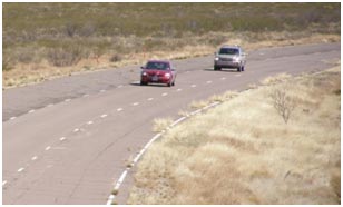 Two vehicles are driving on a test track, one behind the other. The track consists of three paved lanes surrounded by empty grassy fields.