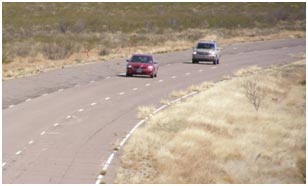 Two vehicles are driving on a test track, one behind the other. The track consists of three paved lanes surrounded by empty grassy fields.