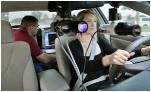 A study participant drives an instrumented vehicle while an experimenter sitting in the backseat monitors the study on a computer. The vehicle includes several recording devices attached to the front windshield.