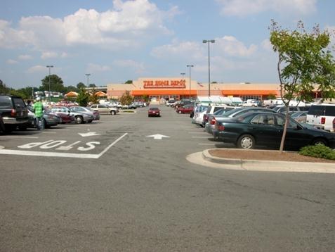 Figure 11. Photo. Example of a parking lot/garage location. This photo shows one row within a parking lot at The Home Depot. The row has an up arrow and down arrow on the pavement in white paint to indicate the direction of travel for motor vehicles. There is a car driving on the right side of the aisle toward the store. The lot appears to be fairy full, and some pedestrians are visible.