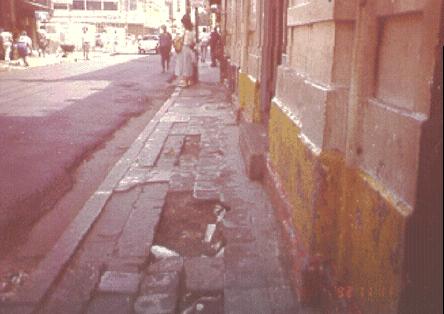 photo of a poor quality pedestrian area with a narrow sidewalk and missing sections of brick in the walkway