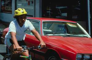 Photo of young boy riding a bicycle on a street past a parked car.