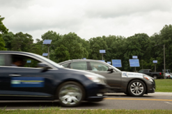 This photograph shows two cars traveling in the same direction on a two-lane road. The car closest to the camera is traveling next to and a few feet behind the car farthest from the camera. In the background, trees and solar panels affixed to poles are shown.