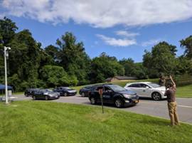 This photograph shows a man standing on the grass next to a roadway. The man has his right arm in the air. On the roadway next to the man, on his right, six vehicles are shown driving on the roadway. Four vehicles are in the lane farthest from the man, and two vehicles are in the lane closest to the man.