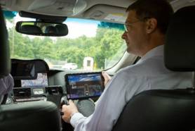 This photograph shows a man sitting in the passenger seat of a car. He is holding a tablet computer with a computerized image of a roadway on it. The man is looking at the driver of the vehicle, who is not shown.
