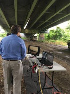 The photograph shows a man standing on the ground under an elevated section of road. A table is shown on the right side of the photograph, and there are two laptops and one computer monitor on top of the table. There are also various wires connected to the laptops and monitor.