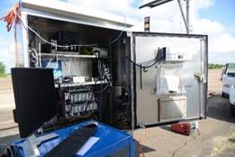 This photograph shows an equipment cabinet with its door open. The cabinet is located on a test track during a traffic simulation. There are many types of electronics and wires in the cabinet, and a wire is hanging on the left side of the cabinet.