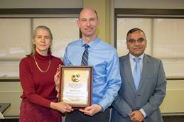 This photograph features three people: two men on the right and a woman on the left. The man in the middle is holding an award. The woman is also holding the award because she is presenting it to the man in the middle.