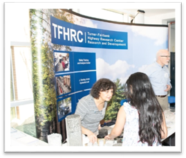 This photograph shows a woman and a man standing behind a table. The woman behind the table is bent slightly forward as she is talking to a young woman, who is on the other side of the table. Both women are located in front of a large poster, and there are various highway–related materials on the table, such as examples of rock, cement, and concrete.