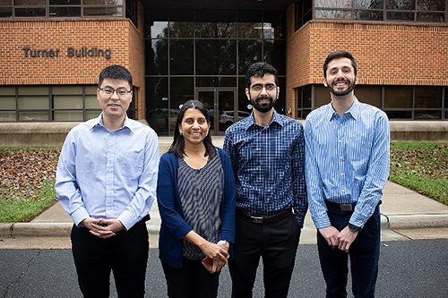 This photo shows four people standing in front of a brick building. From left to right, there is a male figure, a female figure, and two male figures.