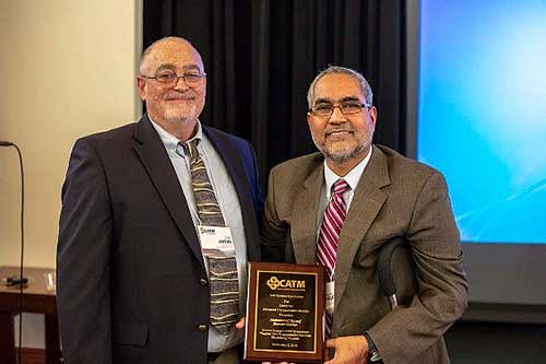 This photograph shows two men in suits standing side by side in a room. The man on the left is holding a wooden plaque. 