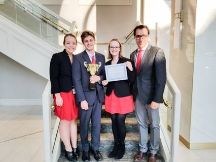 This photograph shows four people standing on a staircase. From left to right, there is a woman, a man holding a trophy, a woman holding a certificate, and another man.