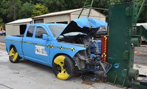 This photograph shows the aftermath of a truck that has crashed into a wall. The front portion of the truck is crumpled and the truck's hood is bent and open.