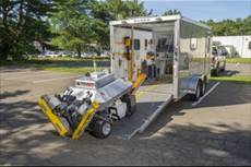 The photograph shows a robot mounted on four wheels disembarking from a trailer.