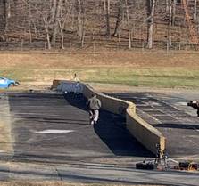 The photograph shows a man walking in front of a long section of a polymer concrete wall. The portion of the wall that was struck by a vehicle is curved inward.