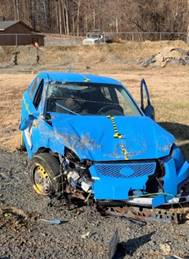This photograph shows a car with its front end smashed, its windshield cracked, and the driver's side door open. The car was involved in a crash test on a closed course.