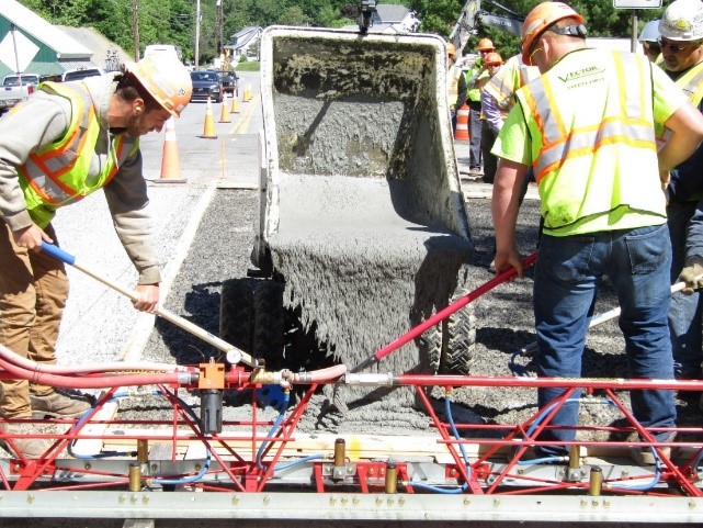 Highway workers use tools to spread out ultra-high performance concrete as it is poured from the back of a truck onto a road surface.
