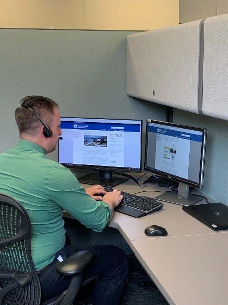 A technical support analyst sits at a desk in front of two computer screens. He is wearing a telephone headset.