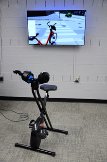 The bicycle simulator is shown in a room facing away from a television screen. The television screen shows an image of what the user of the bicycle simulator would see while wearing the virtual reality headset.