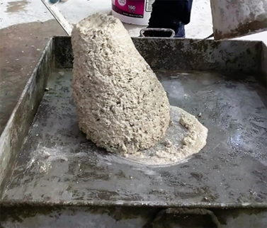 Photograph of a slump test specimen performed on stiff, low-slump, freshly mixed concrete made with alternative magnesium phosphate cement. The slump specimen is in a metal mixing pan. The mixture is a light gray color and is tipping to the left.