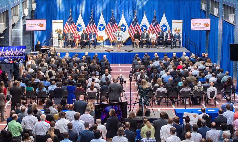 A room with a seated and standing audience is shown. The audience is seated in front of a dais, and a woman is giving a speech. Eleven people are seated on the dais.