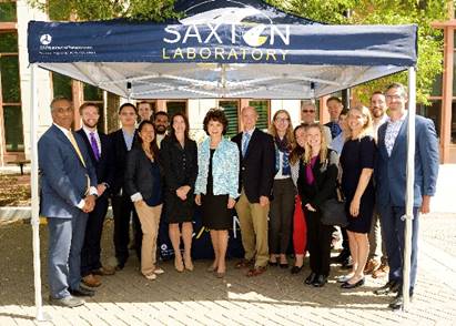 Eighteen people are photographed outside under a tent supported by four poles.